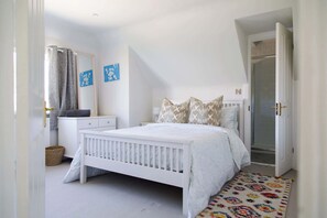A bedroom with a white crib, a bedside table, a mirror, and a door leading to a bathroom. - Beach house with pool in West Wittering (West Wittering)
