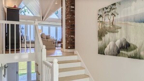 Een ruime woonkamer met een trap naar een hoger gelegen verdieping, een groot raam en een schilderij van bomen aan de muur.