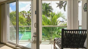 Een kamer met een schuifglazen deur die uitkomt op een balkon met uitzicht op een tennisbaan en palmbomen.