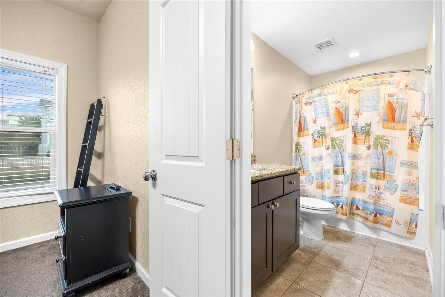 A bathroom with a toilet, a shower curtain with a beach theme, a black cabinet, and a window with blinds.