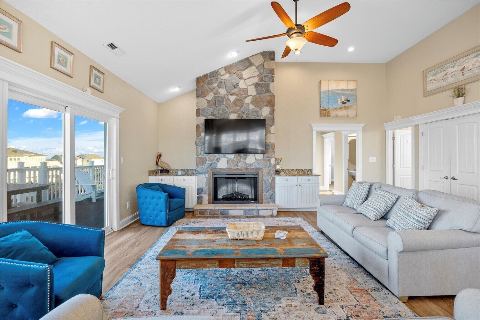 A spacious living room with a stone fireplace, a ceiling fan, and a view of the outside.