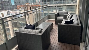 Ein Balkon mit Korbmöbeln und Glastrennwand, mit Blick auf eine Skyline mit Hochhäusern.
