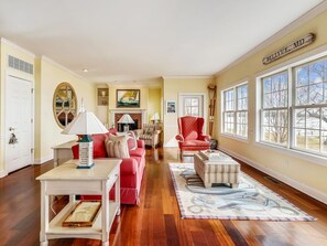 Un salón amplio con pisos de madera, una chimenea, un sillón rojo y ventanas grandes que ofrecen vistas al exterior.