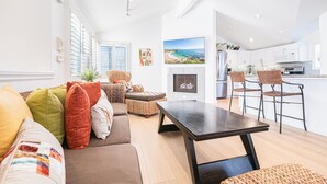 Una sala de estar moderna con un sofá, una mesa de centro y sillas de mimbre. Hay una chimenea y una zona de cocina con taburetes de bar.