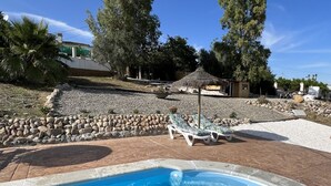 En swimmingpool med en kant av sten, solstolar och en byggnad med halmtak, allt placerat i ett välordnat område med träd och en byggnad i bakgrunden.