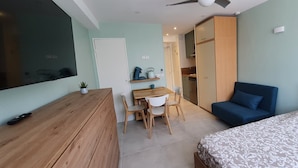 A compact bedroom with a bed, a desk with chairs, a wardrobe, and a kitchenette in the background. - Comfort studio - Double bed + single bed - FAME MARINA BAIE DES ANGES (Villeneuve-Loubet)