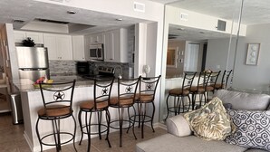 A modern kitchen with white cabinets, a stainless steel refrigerator, and a microwave. There are four bar stools with star-shaped cutouts and a grey sofa with patterned cushions.