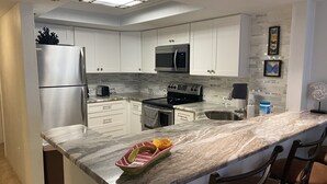 A modern kitchen with white cabinets, a stainless steel refrigerator, a microwave, and an oven. There is a marble countertop with a small dish and a towel.