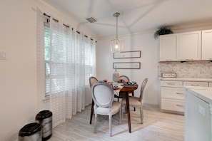 A dining area with a table set for two, chairs, a pendant light, and a kitchen with white cabinets and a tiled backsplash.