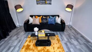 A living room with a black sofa, two floor lamps, a glass coffee table, and a painting on the wall.