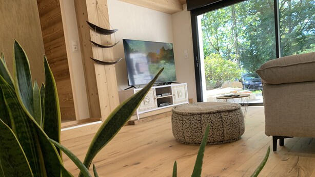 A modern living room with a wooden floor, a round ottoman, a television, and large windows offering a view of greenery outside.