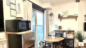 Cocina compacta con refrigerador, microondas y tostadora. Hay un comedor con mesa y sillas de madera, y un ventanal con vista a la ciudad.