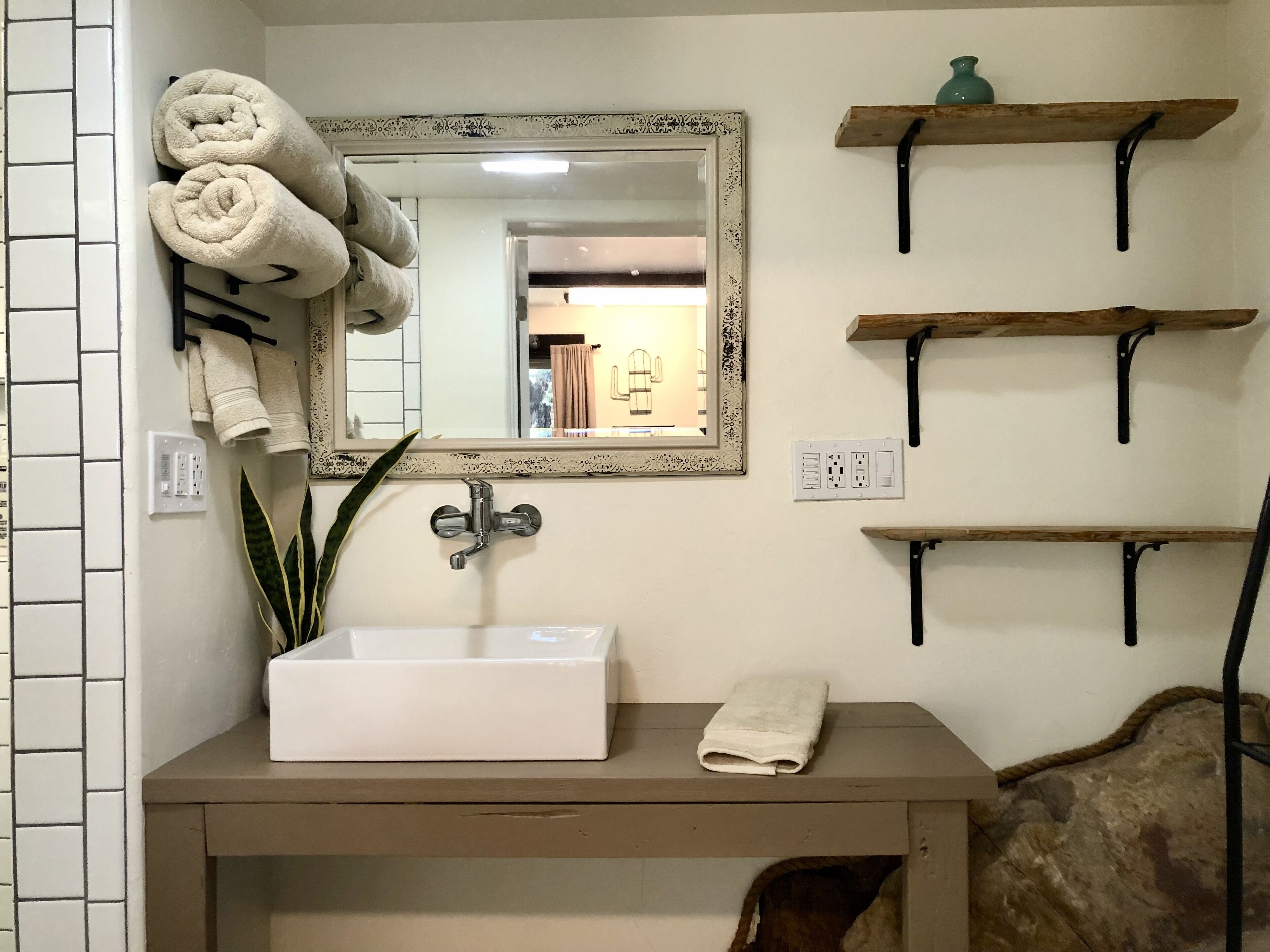 Un baño con un lavamanos blanco, un espejo con marco decorativo y tres estantes de madera.