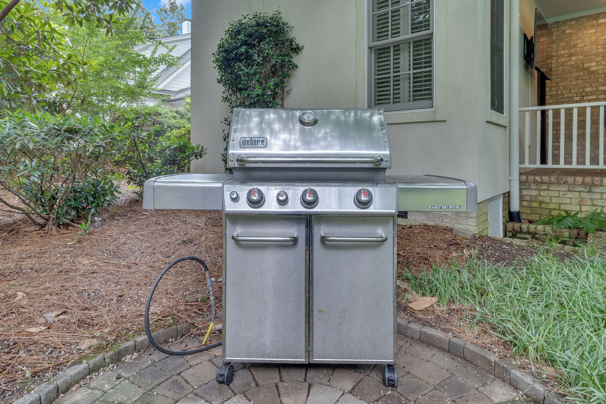 En sølvfarvet Weber Genesis transportabel gasgrill med fire brændere, en sideskuffe og en skorstensstarter, placeret på en belagt flade med et murstenshus i baggrunden.