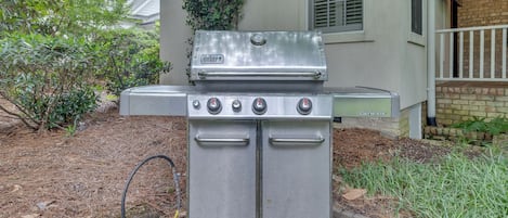 En sølvfarvet Weber Genesis transportabel gasgrill med fire brændere, en sideskuffe og en skorstensstarter, placeret på en belagt flade med et murstenshus i baggrunden.