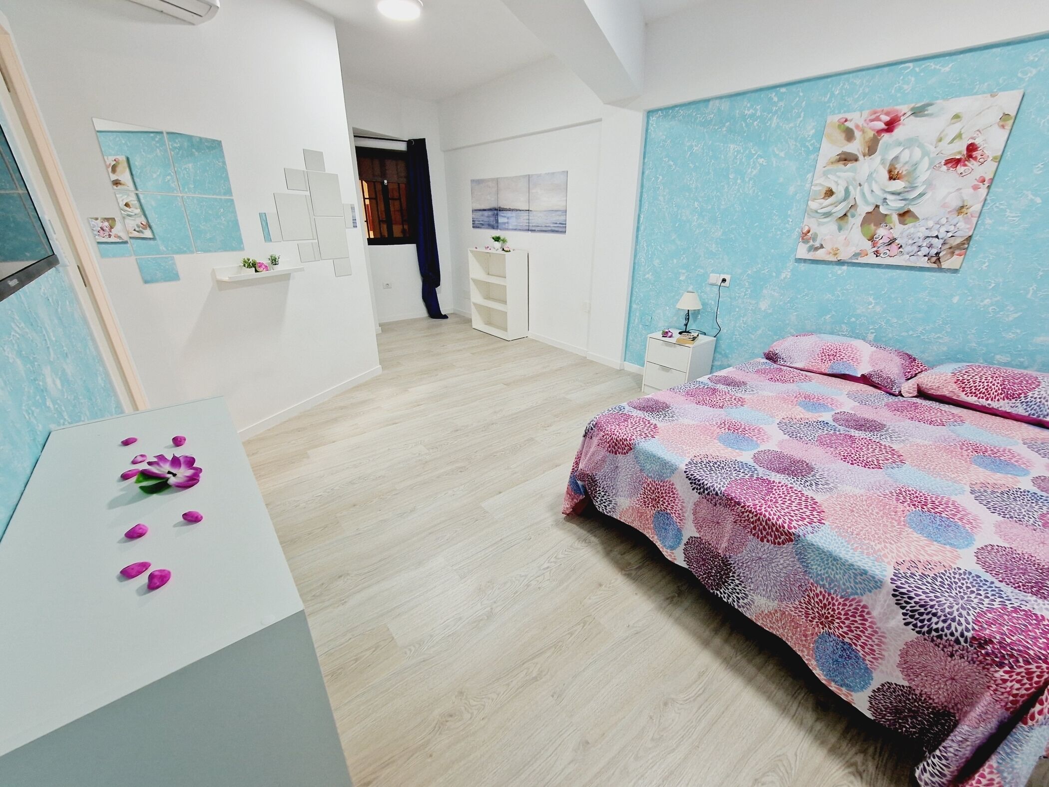 A bedroom with a bed, a desk, and a nightstand. There is a window with curtains, a wall with a floral painting, and a door leading to another room.