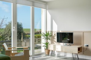 A modern living room with a large sliding glass door, a wooden chair, a TV stand, and a potted plant. - Beach Life, Cabin Vibes, Ocean Views nr Bude (Bude)