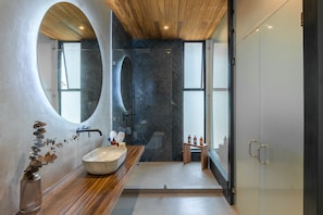 Features a unique oval mirror, large sink, and minimalist design with a wooden vanity and glass shower enclosure.