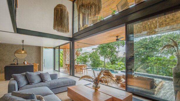 Una sala de estar moderna con un sofá seccional, una mesa de centro de madera y ventanas amplias que dan a una piscina y follaje.