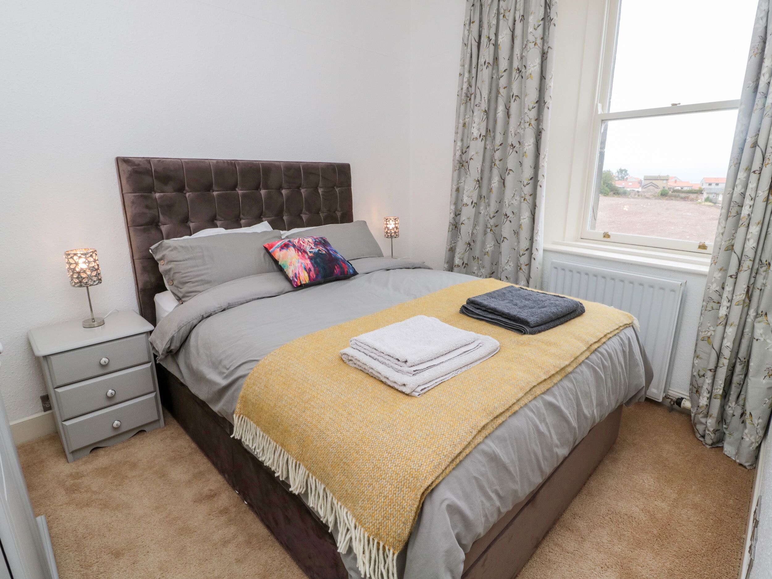 A neatly made bed with a headboard, a bedside table with a lamp, and a window with curtains.