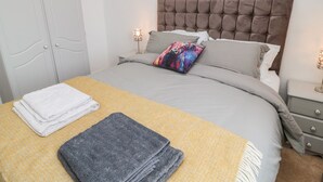 Een netjes opgemaakt bed met een grijze en gele sprei, een grijze plaid en een kleurrijk kussen. Er is een witte deur en een nachtkastje met een lamp.