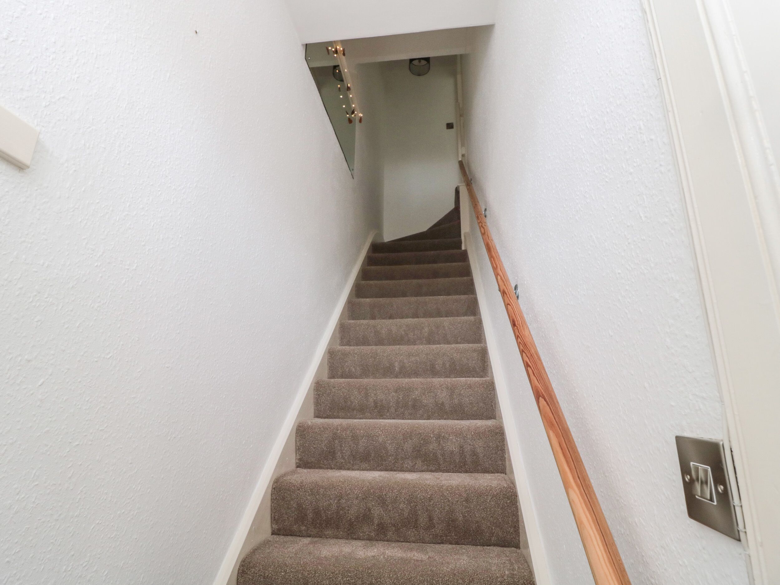 Un escalier recouvert de moquette, avec des rampes en bois, des murs blancs et un interrupteur sur le mur.