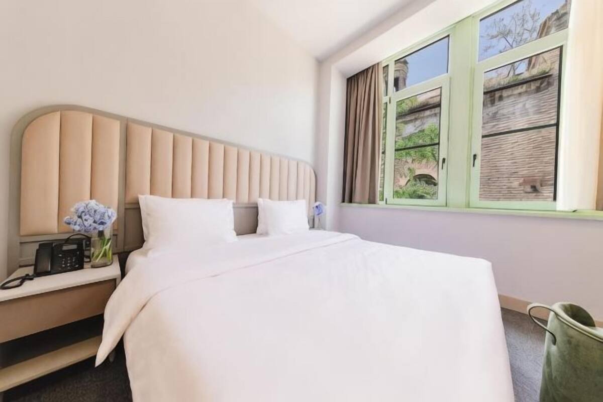 Una camera d'albergo con un letto, un comodino con un telefono, un vaso con fiori e una finestra vista verde.
