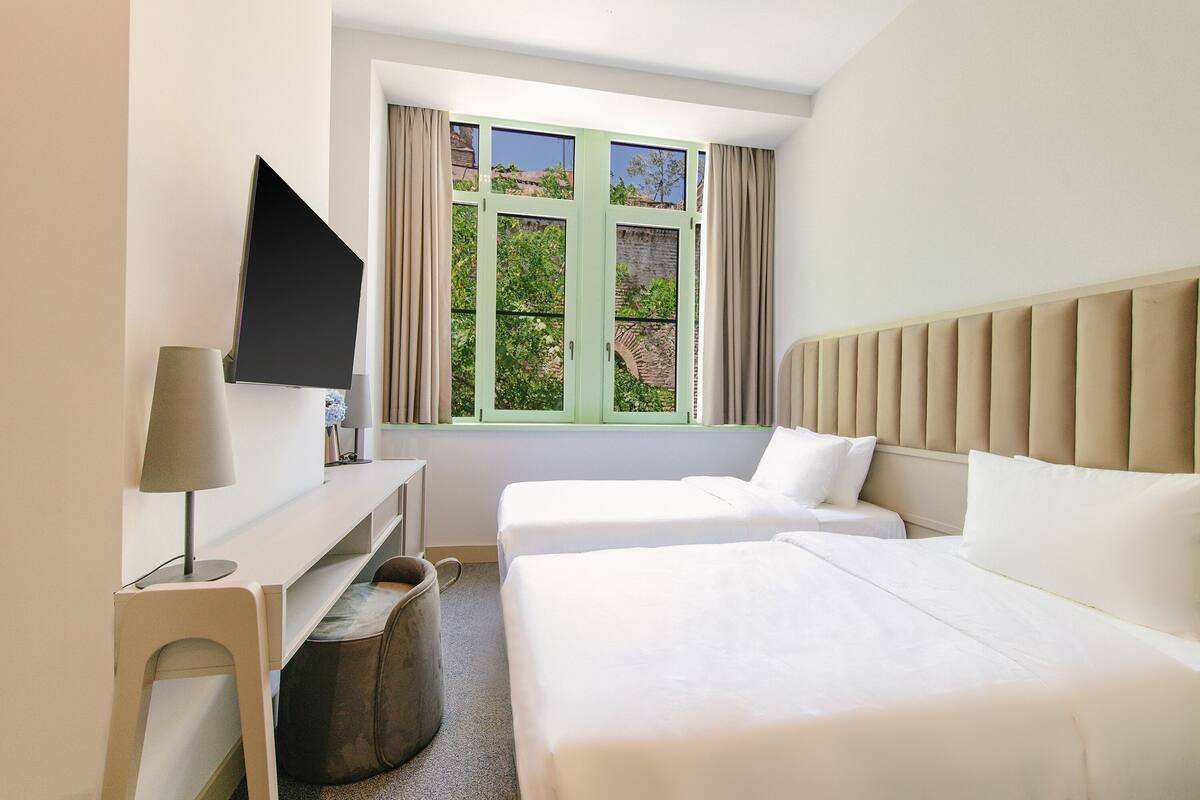 Una camera d'albergo con due letti, una scrivania, una televisione e una finestra con tende.