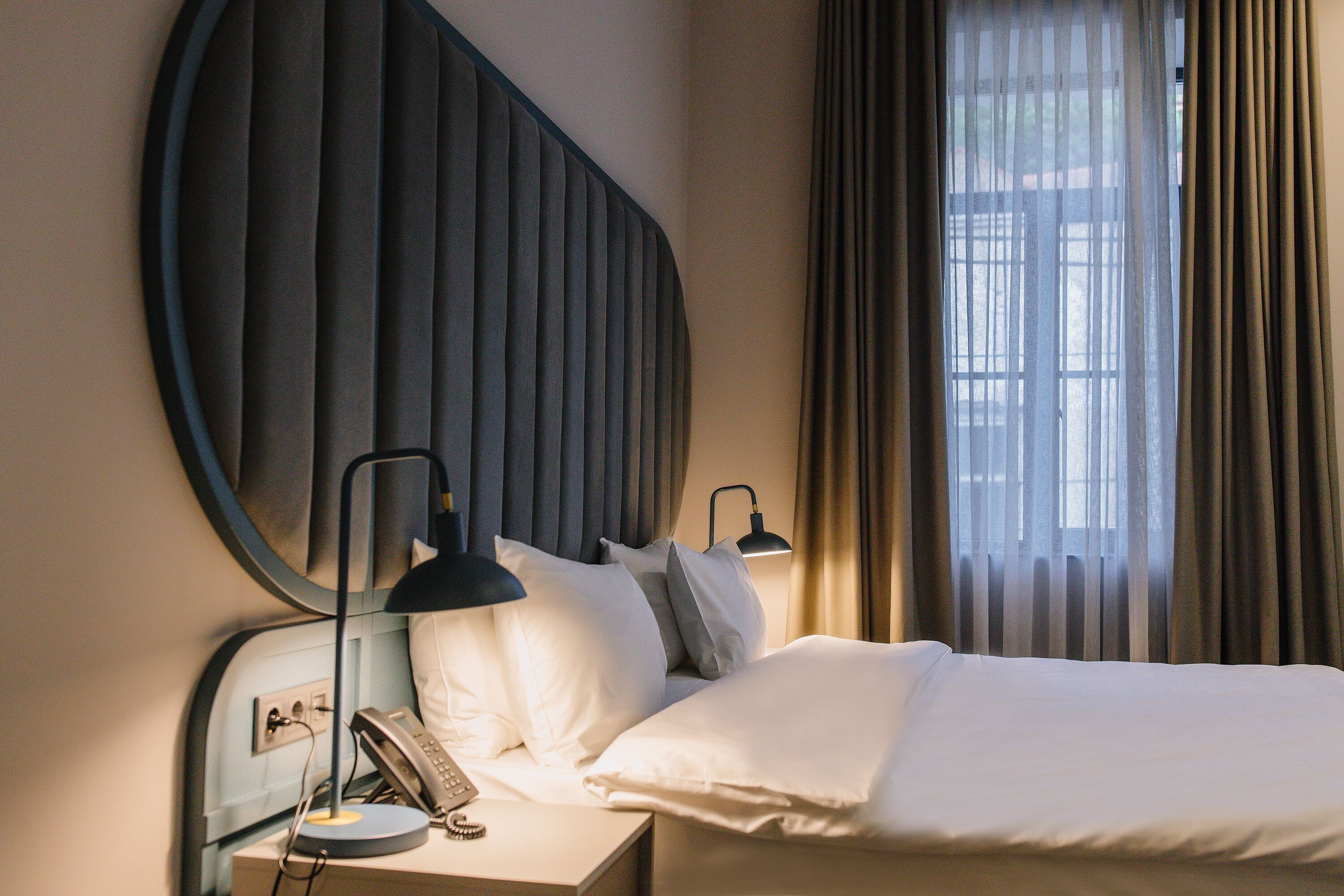 A hotel room with a bed, bedside lamps, a telephone, and a window with curtains.
