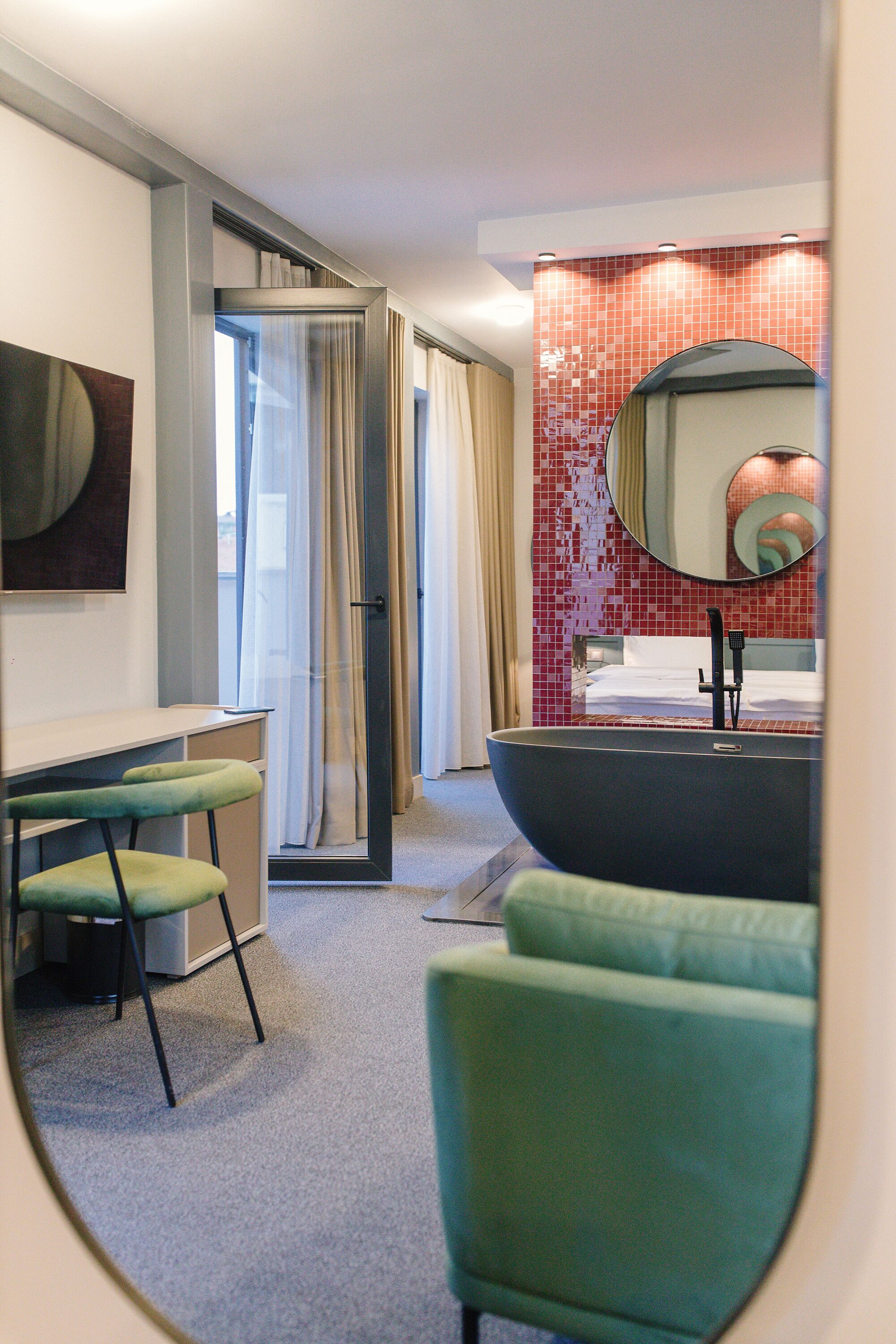 Une salle de bain d’hôtel avec une baignoire, un lavabo et un miroir. Il y a une porte qui donne sur une autre pièce et une fenêtre avec des rideaux.