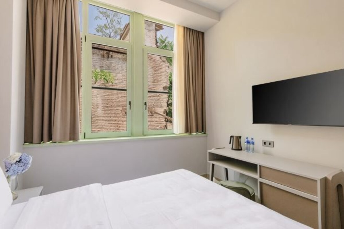 A hotel room with a bed, a desk with a flat-screen TV, a window with curtains, and a brick wall outside.