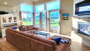 A spacious living room with a large brown sectional sofa, a flat-screen TV mounted on a stone wall, and a view of a mountainous landscape through large windows. - New home in the heart of Alpine! (Alpine)