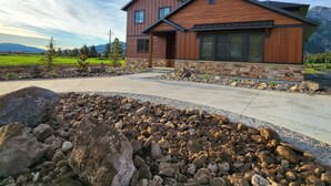 A modern two-story house with a stone foundation and wooden siding, surrounded by a landscaped garden with large rocks and a view of mountains in the background. - New home in the heart of Alpine! (Alpine)