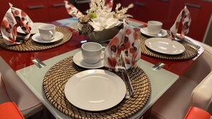Une table à manger dressée avec de l’assiette, des bols et des couverts blancs, entourée de sets de table en fibres tressées et de chaises rouges.