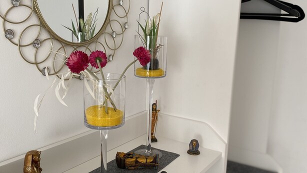 Une coiffeuse blanche avec un miroir, deux vases en verre ornés de fleurs, une petite statue et un cadre métallique décoratif accroché au mur.