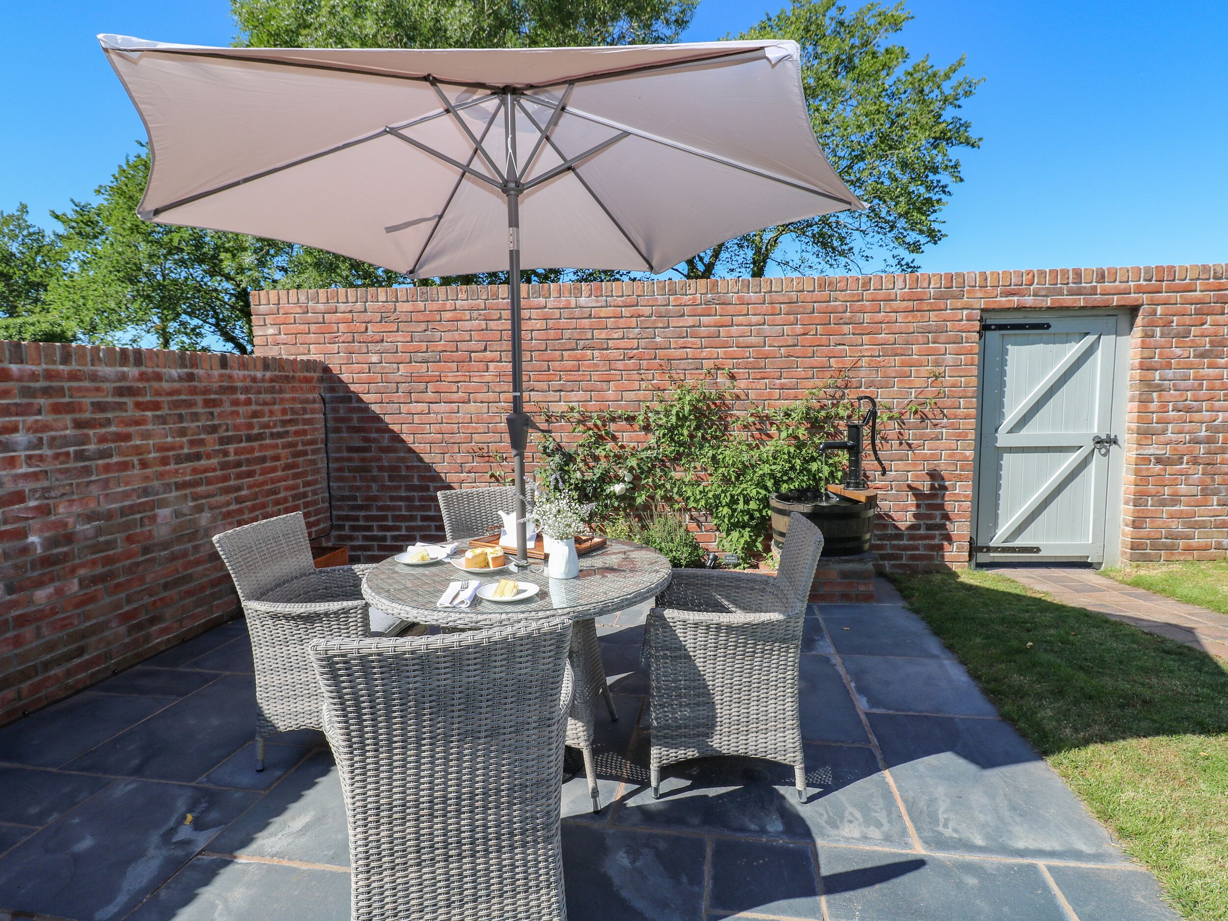 Une terrasse dotée de meubles en osier, d’une table dressée avec des assiettes et une carafe, et d’un grand parasol.
