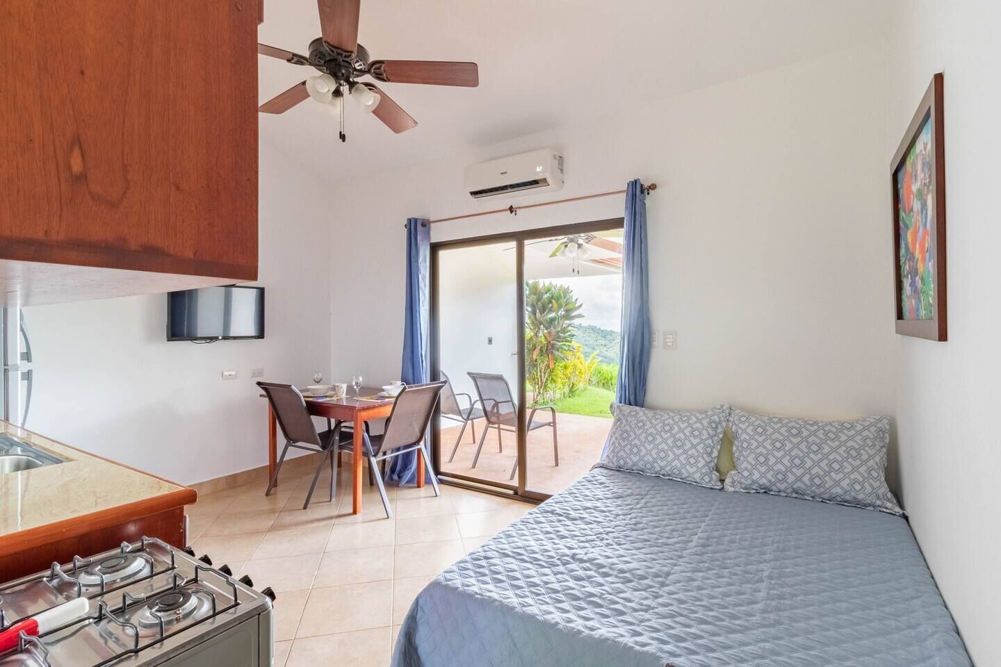 Una habitación con cama, una mesa de comedor con sillas, una zona de cocina con estufa, un ventilador de techo y vistas a través de una puerta corrediza de vidrio.