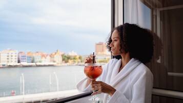 Iemand in een witte badjas met een cocktailglas bij een raam met uitzicht op een haven, met gebouwen op de achtergrond.