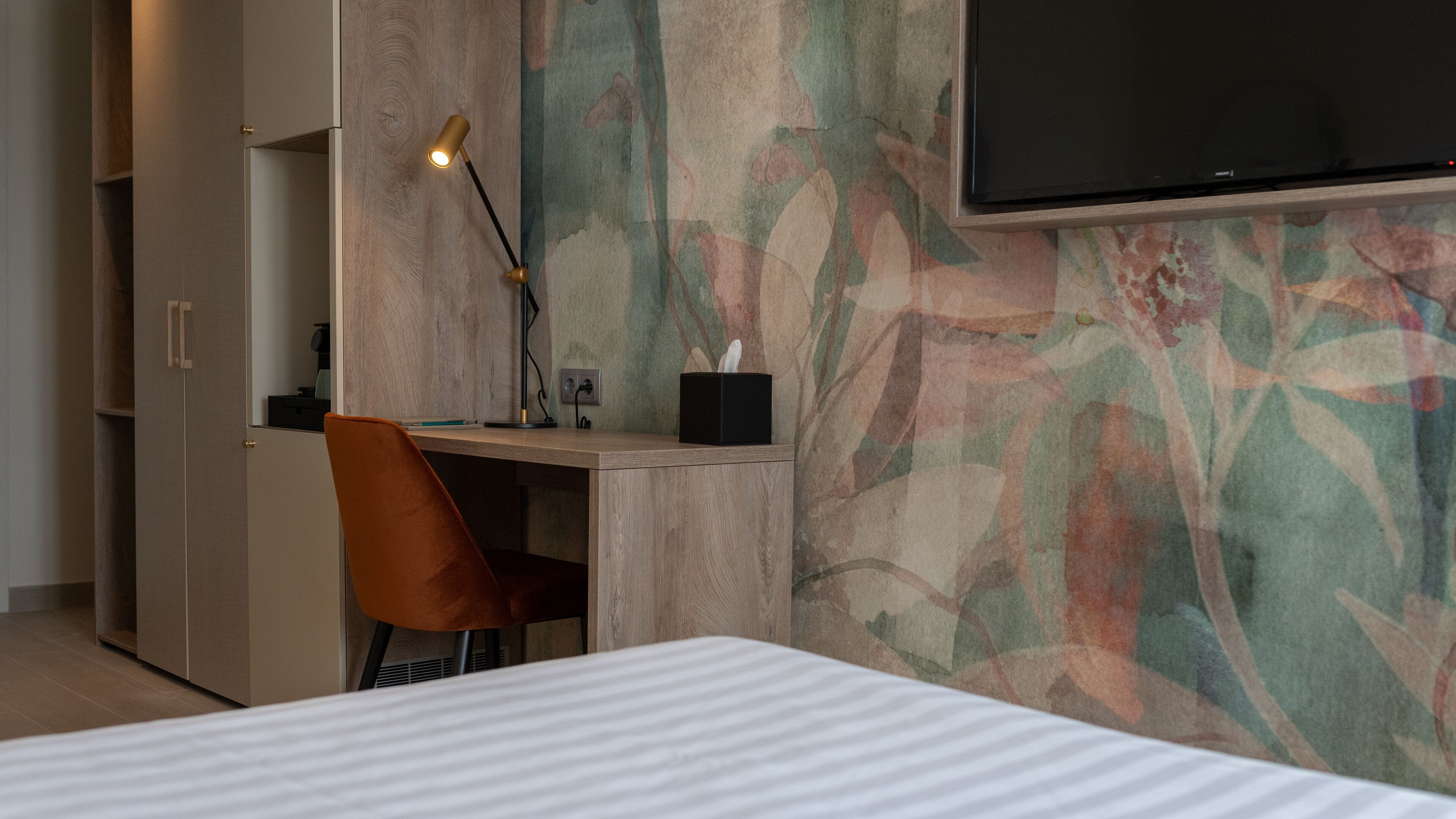 A hotel room with a bed, a desk with a lamp, a chair, a TV mounted on the wall, and a wardrobe.