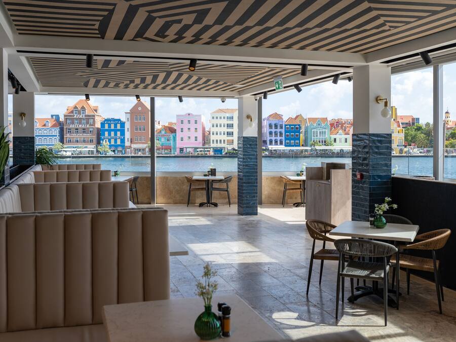 A restaurant with a terrace overlooking a waterfront, featuring colorful buildings in the background.