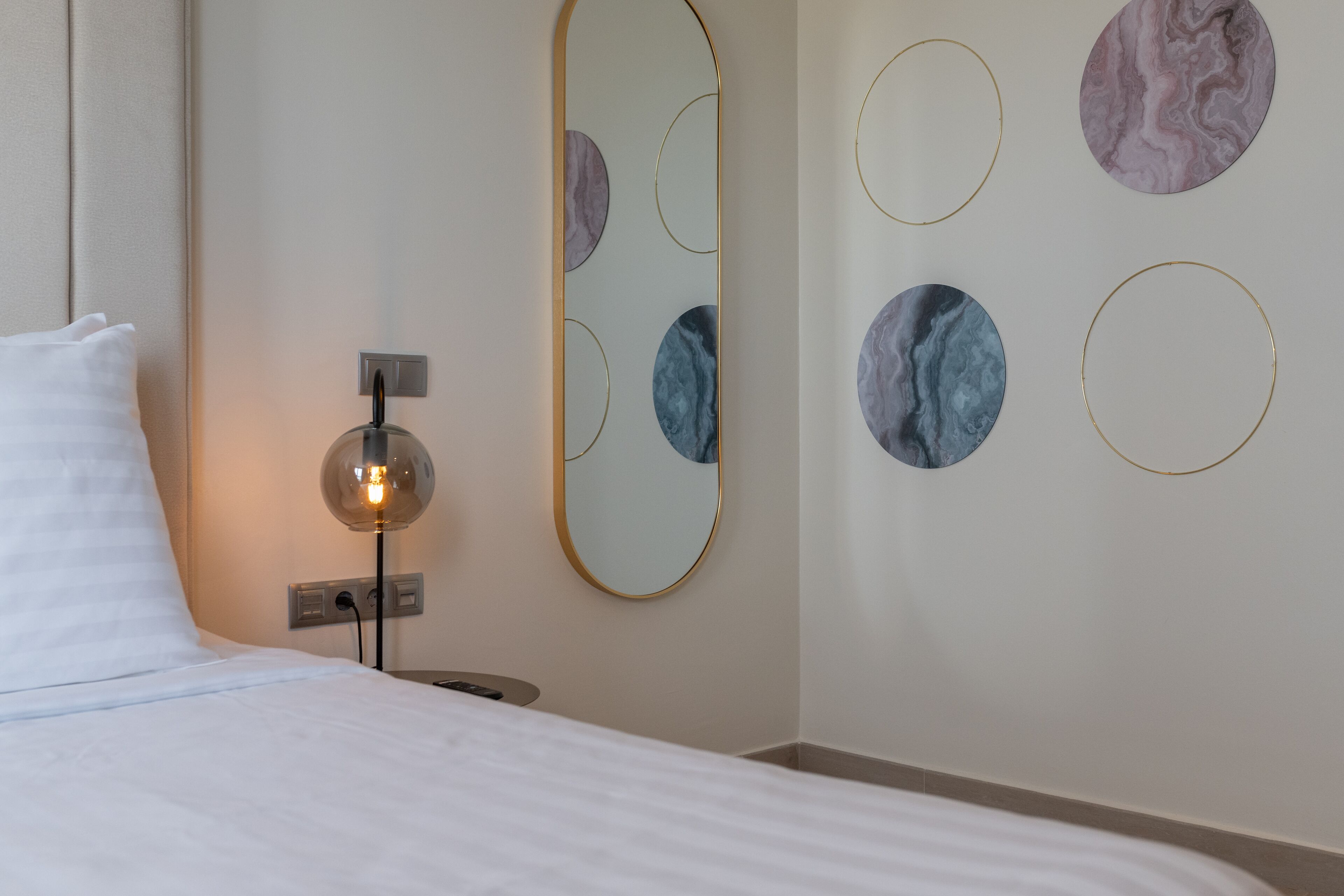 A hotel room with a bed, a bedside lamp, a wall-mounted mirror, and circular wall art.