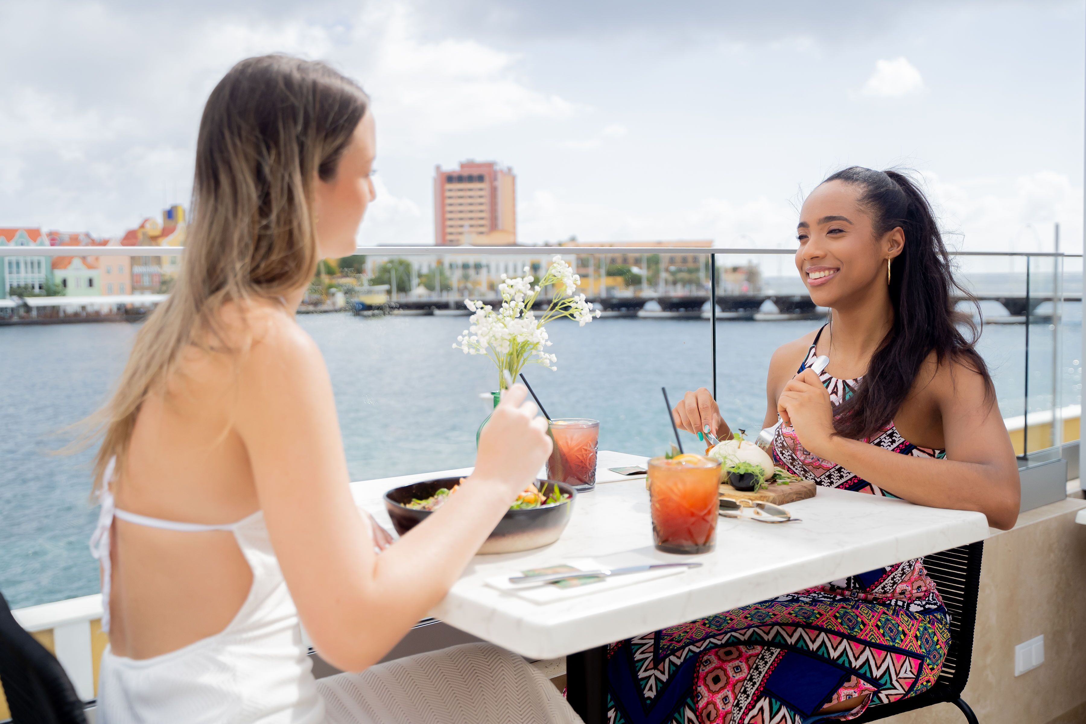 Twee personen dineren aan een tafel met uitzicht op een wateroppervlak en gebouwen op de achtergrond.