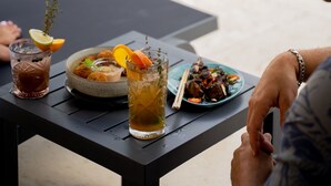 Una mesa de comedor con comida y bebidas, una piscina al fondo y una persona sentada junto a la piscina.