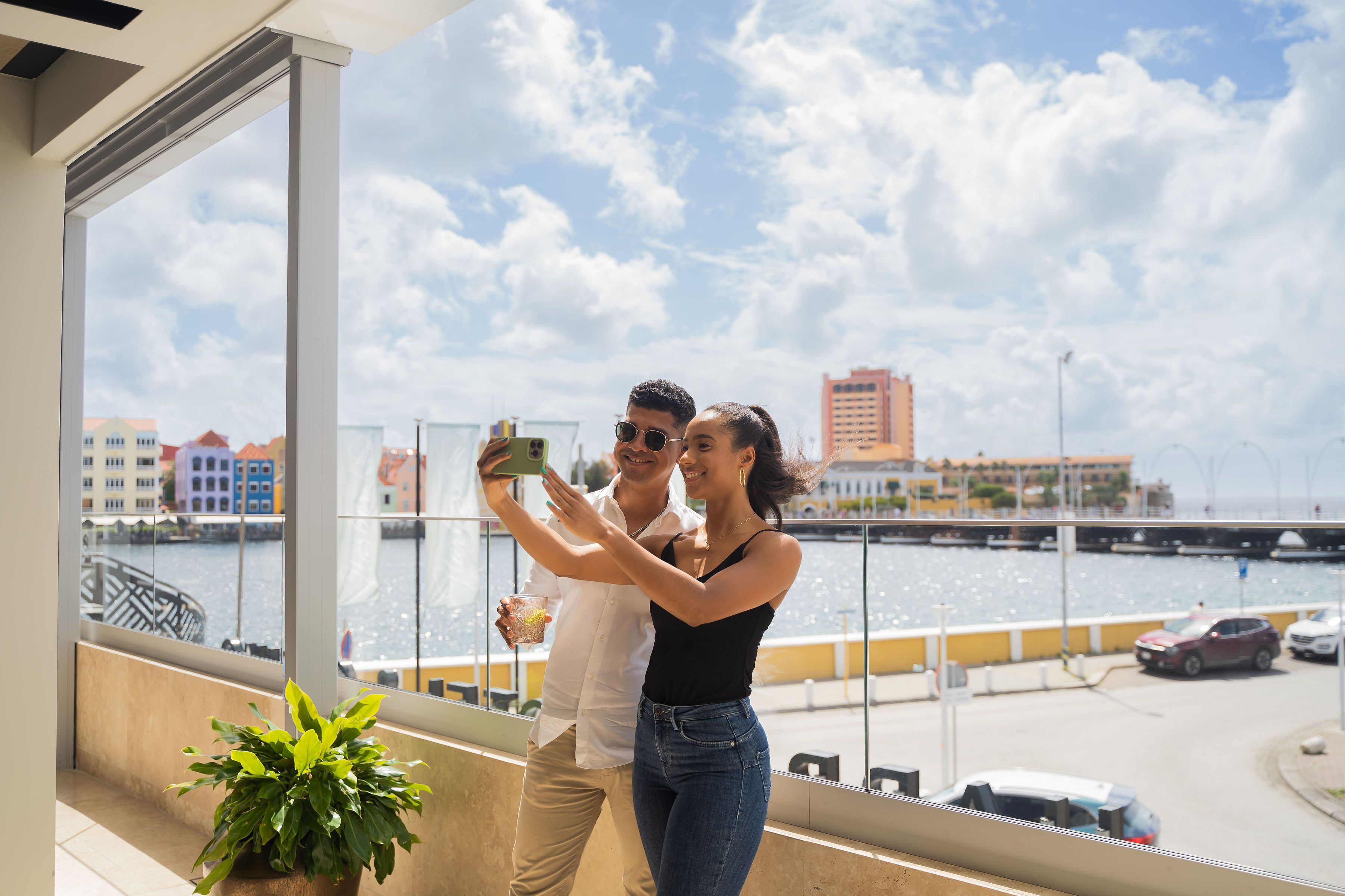 A couple taking a selfie on a balcony with a view of a waterfront and buildings in the background.