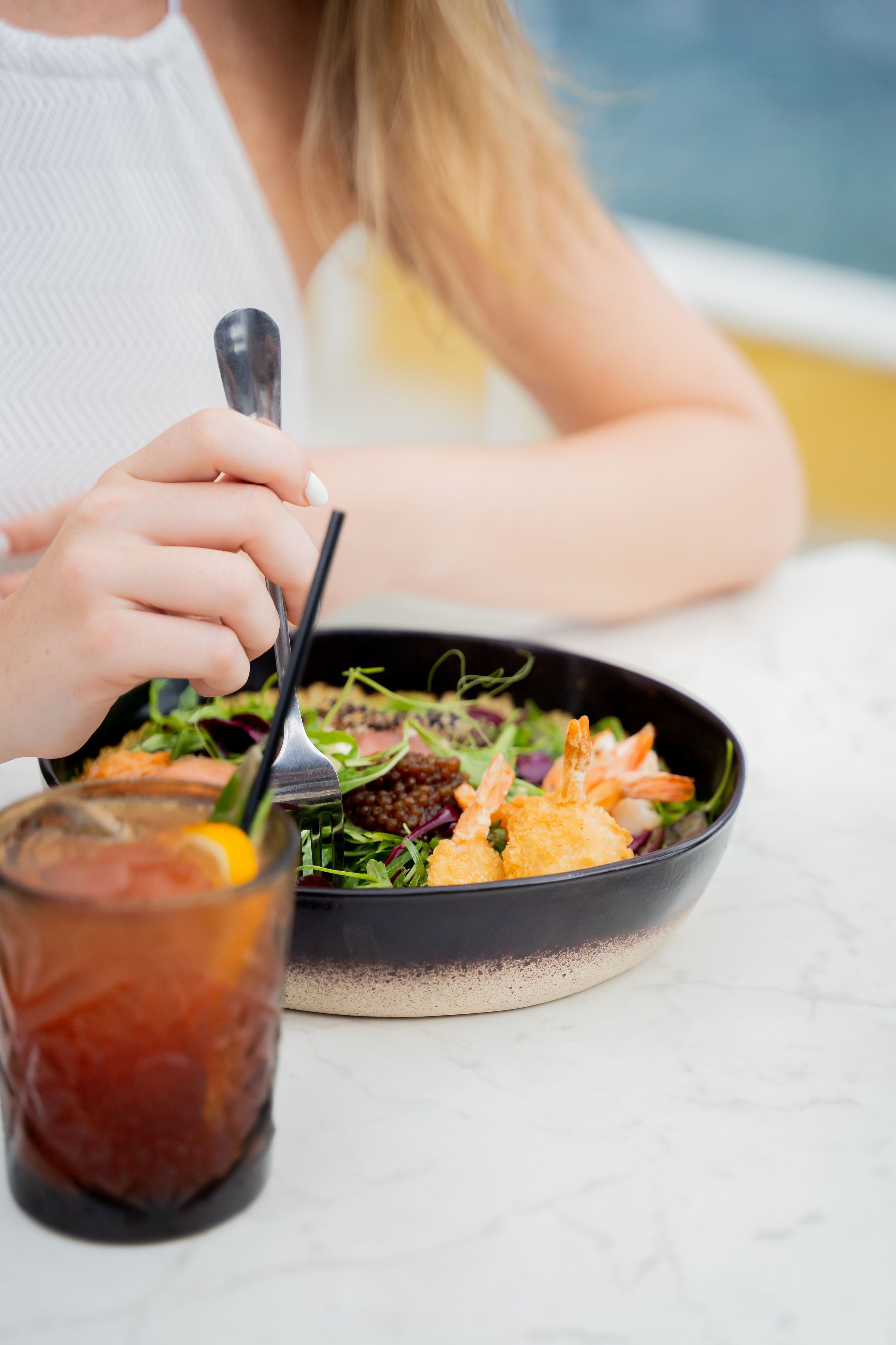 Eine Person isst an einem Tisch. Auf dem Tisch steht eine schwarze Schüssel mit einem Salat und ein Glas Eistee.