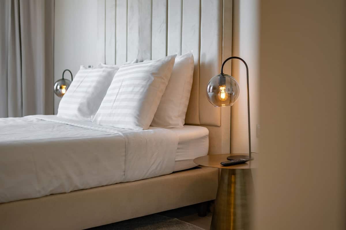 Una cama bien tendida con sábanas blancas, una mesita de noche con un control remoto y una lámpara de pared.