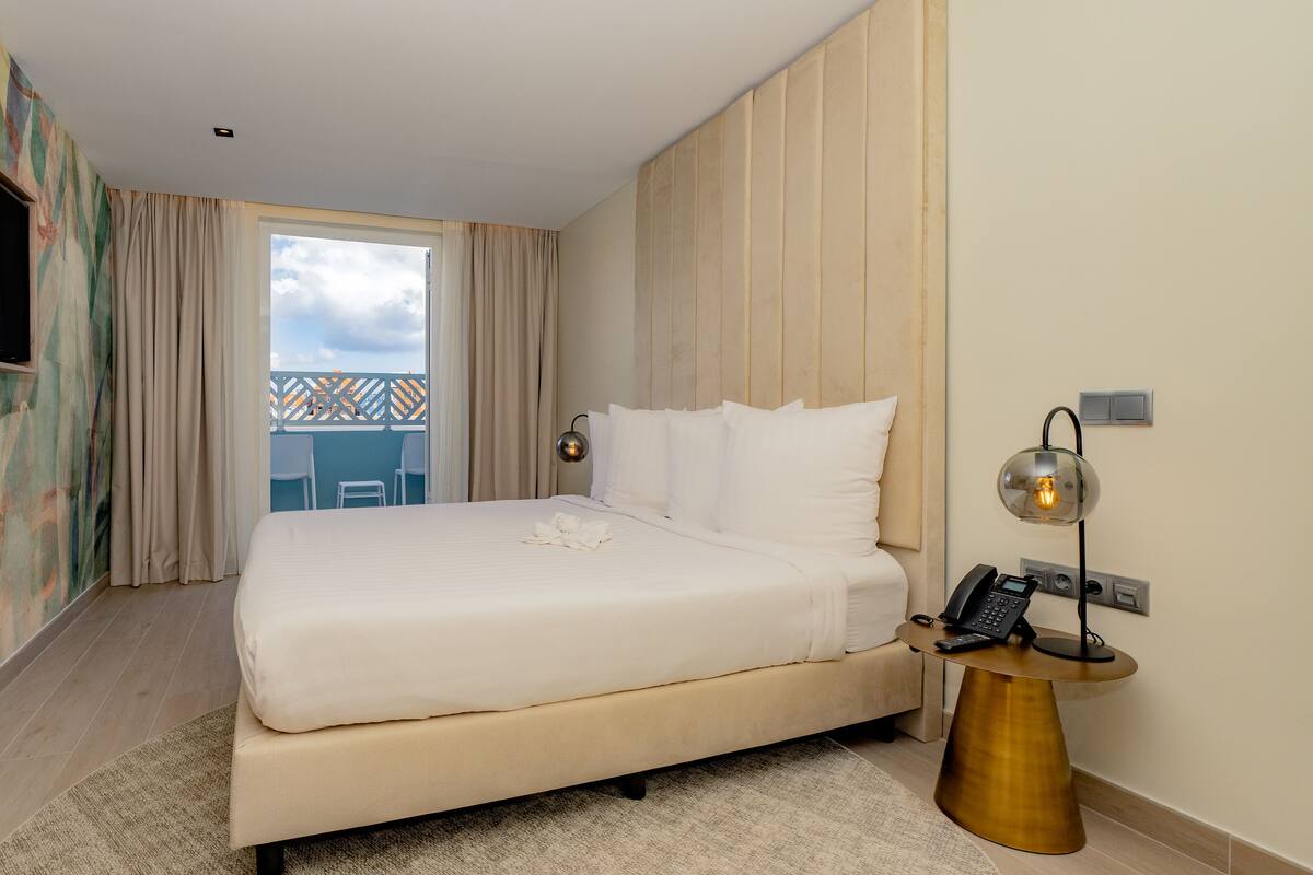 Habitación de hotel con una cama, un televisor montado en la pared, una mesita de noche con una lámpara y vistas al balcón exterior.