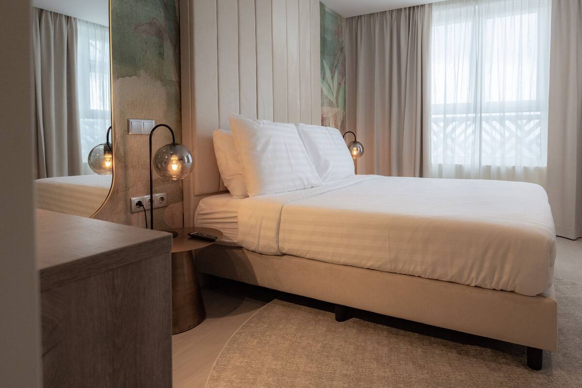 Una habitación de hotel moderna con una cama grande, dos lámparas de noche, una mesita de noche y vistas al exterior a través de cortinas traslúcidas.