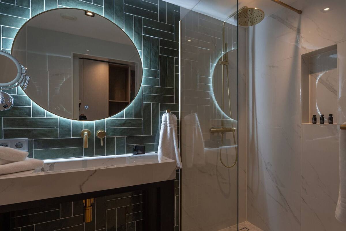 Un baño moderno con ducha envuelta en vidrio, un espejo redondo y un tocador con encimera de mármol.