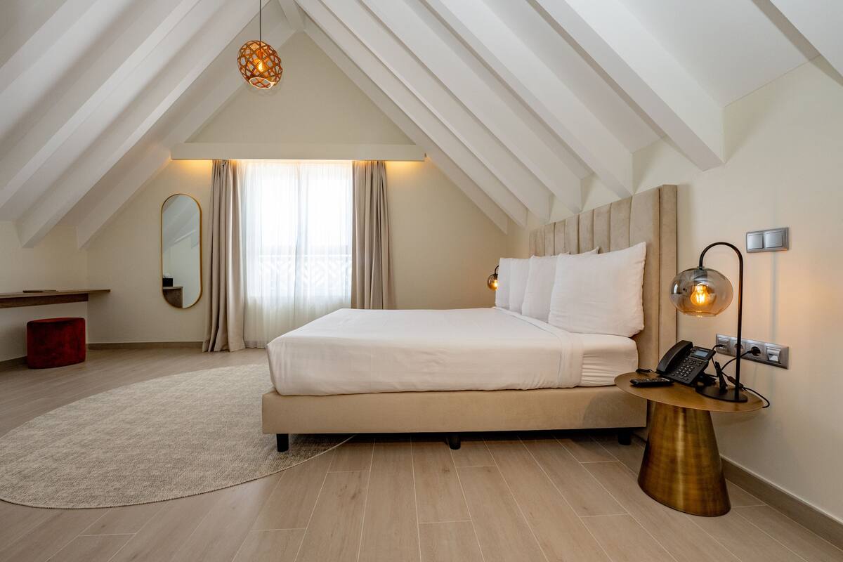 Un dormitorio con una cama grande, una mesita de noche con lámpara, un espejo y un otoman rojo.